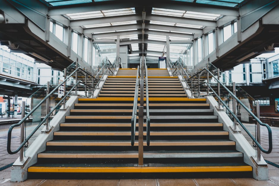 A wide view of an outdoor staircase leading up to a modern transparent glass and metal pedestrian footbridge at a bus or train station. The staircase features multiple steps with black treads and yell